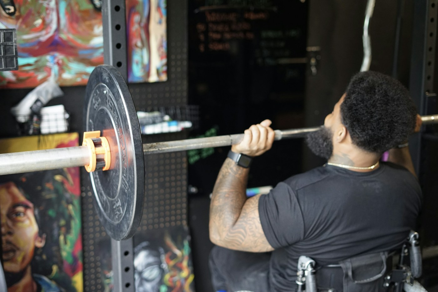 A Black athlete practising a barbell lift from his wheelchair in a gym
